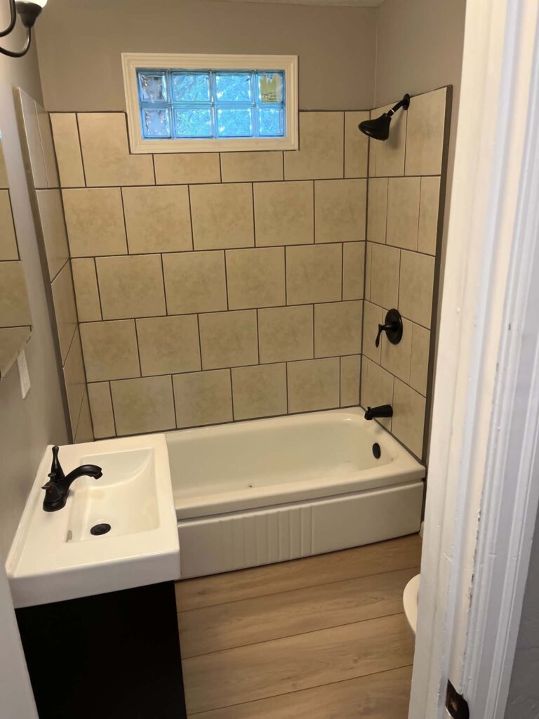 Clean tub and shower with tan tile, glass-block window, and black fixtures