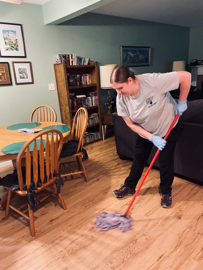 Cleaner in a gray shirt and gloves mopping a hardwood floor beside a dining table