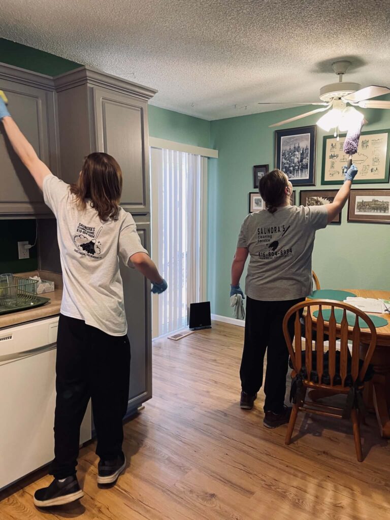 Two cleaners dusting kitchen cabinets and wall art in a green kitchen